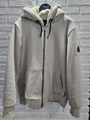 Moose Knuckles CLASSIC BUNNY hoodie/jacka  - Säljer en Moose Knuckles CLASSIC BUNNY beige hoodie/jacka med dragkedja och stor huva med fluffigt foder. Den har svarta snören, två stora fickor fram och en patch på ärmen. Perfekt för dig som gillar en clean och stilren look. Storlek XL men passar även dig som har L men gillar ha lite baggy. Använd få tal gånger och inga täcken på användning. Köpt på johnells och kvitto finns digital om du vill ha.