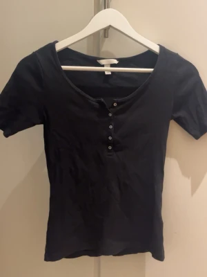 Svart t-shirt med knappar - En enkel svart t-shirt med korta ärmar och rundad halsringning. Säljer denna eftersom det inte är min stil och den har aldrig riktigt kommit till användning. Passar till jeans och stickad tröja men också till kjol.