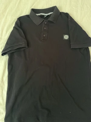 Svart pikétröja från Stone Island - Klassisk svart pikétröja från Stone Island med krage och tre knappar framtill. Tröjan har korta ärmar och den ikoniska Stone Island-loggan på bröstet. Perfekt till en clean och stilren look.