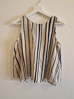 Randig ärmlös topp med öppen rygg - Säljer en vit ärmlös topp med svarta vertikala ränder. Toppen har en lös passform och en snygg öppen detalj i ryggen. Perfekt för sommaren och enkel att matcha med jeans eller shorts. Materialet känns lätt och luftigt.