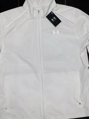 Vit vindjacka från Under Armour - Snygg vit vindjacka från Under Armour med dragkedja framtill och hög krage. Jackan har två sidofickor, reflekterande detaljer och logga på bröstet samt text på ryggen. Tillverkad i lätt och vindtätt material, perfekt för aktiva dagar.
