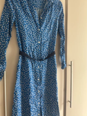 Blommig blå klänning Tommy Hilfiger - Snygg blå klänning från Tommy Hilfiger med vitt blommönster, lång ärm och knappar framtill. Klänningen har ett bälte i midjan och klassisk krage. Perfekt för dig som gillar en feminin och fräsch stil.