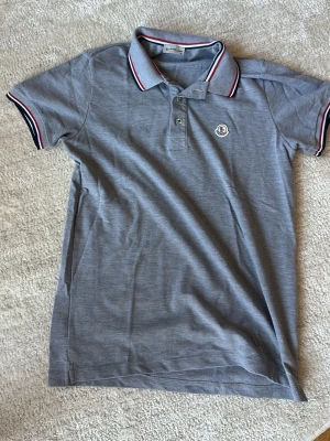 Grå pikétröja från Moncler - Snygg grå pikétröja från Moncler med klassisk krage och knäppning framtill. Tröjan har korta ärmar och detaljer i rött, vitt och blått på krage och ärmslut. Moncler-logga på bröstet. Perfekt för en stilren look.