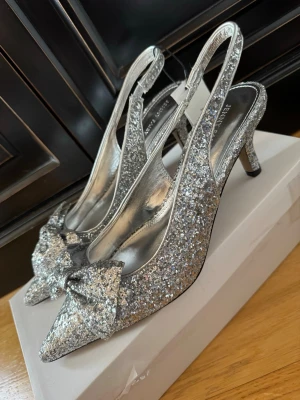 Glittriga silverpumps med rosett - Nya snygga klackskor / klackar i silverglittrigt material, från JENNY FAIRY. Otroligt fina. Spetsig tå, öppen häl med rem och en rosett framtill.  Snygga till balklänningen och / eller sommarens finkläder. Storlek 40. Lapparna kvar. Finns inget att anmärka på. NWT. 