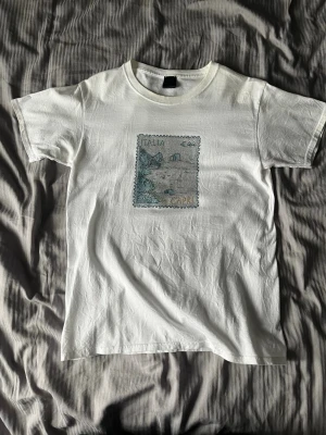 Vit Bvaldi t-shirt - Vit t-shirt från Bvaldi med ett unikt tryck i blått och beige på bröstet. Klassisk passform med rund hals och korta ärmar. Tillverkad i mjuk bomull som känns skön mot huden. Perfekt för dig som gillar stilrena plagg med en twist.