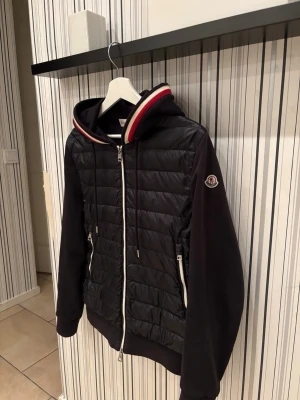 Svart cardigan från Moncler - Snygg svart cardigan från Moncler med quiltad front, hel dragkedja och två dragkedjefickor. Jackan har huva med röd och vit randig kant samt Moncler-logga på ärmen. Perfekt för kalla dagar och riktigt trendig stil.