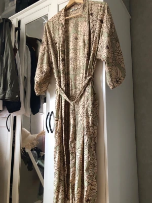 Blommönstrad kimono/morgonrock i viskos - Snygg kimono-inspirerad morgonrock i ljusgrönt viskostyg med stora beige blommor och bruna konturer. Vida ärmar och knytskärp i midjan. Perfekt för chill dagar hemma och ger en avslappnad men ändå stilren vibe. Stl S/M