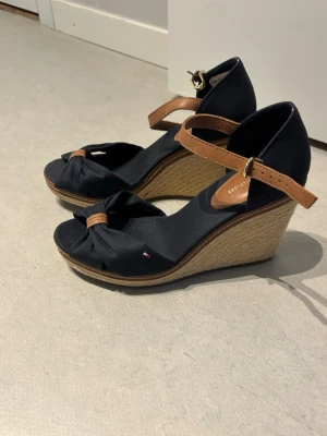 Svarta kilklackssandaler Tommy Hilfiger - Snygga svarta sandaler med kilklack från Tommy Hilfiger. Skorna har bruna remmar i läderimitation, öppen tå med knutdetalj och flätad sula i repmaterial. Perfekta för en trendig och avslappnad look.