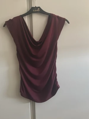 Vinröd draperad topp med djup ringning - Snygg vinröd topp med draperad front och djup ringning både fram och bak. Ärmlös modell i ett mjukt, glansigt material som ger ett elegant fall. Perfekt för dig som vill sticka ut med en stilren och trendig look.