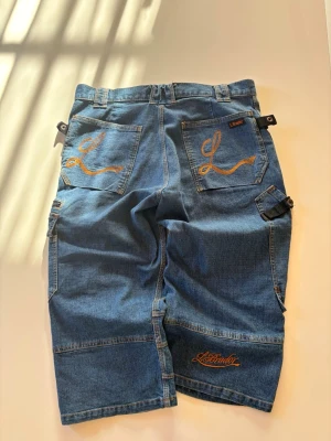Blå jeansshorts från L.Brader - Säljer ett par blå jeansshorts från L.Brader med flera fickor och orange broderier på bakfickorna.
