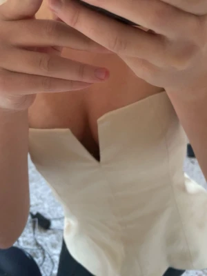 Krämvit korsettopp med unik urringning - Säljer en krämvit korsettopp med snygg, kantig urringning. Toppen är figurnära och har en stilren, minimalistisk look som passar perfekt till jeans eller kjol. Materialet känns slätt och har en lätt glans. Perfekt för dig som vill sticka ut lite extra.