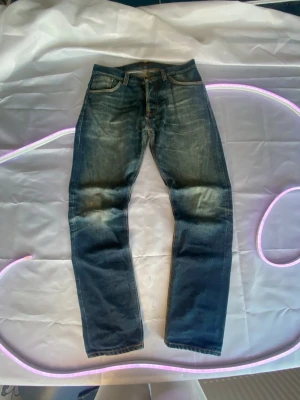 Blå jeans från Nudie Jeans fearless Freddie - Säljer ett par klassiska blå jeans från Nudie Jeans med fem fickor och orangea kontrastsömmar. Jeansen har en normal passform och är tillverkade i bomull. Perfekta för dig som gillar en tidlös och avslappnad stil.
