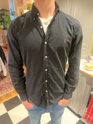 Svart skjorta från Ralph Lauren - Klassisk svart skjorta från Ralph Lauren med knappslå och vit knappstängning. Skjortan har lång ärm och en liten lila broderad logga på bröstet. Tillverkad i mjuk bomull och har en normal passform. Perfekt till jeans eller chinos.