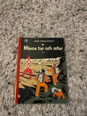 Tintins äventyr: Månen tur och retur - Säljer 'Månen tur och retur' del 2 från serien Tintins äventyr av Hergé. Omslaget är färgstarkt med astronauter i orange dräkter och en rödvit raket på månen. Boken har hårda pärmar med röd rygg och klassiska tecknade illustrationer.
