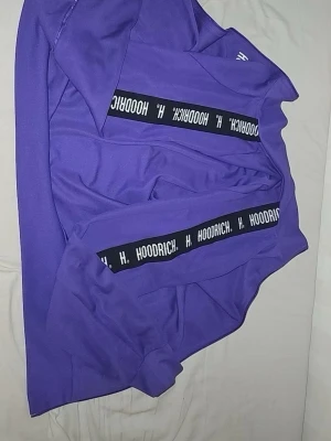 Lila hoodie från Hoodrich - Snygg lila hoodie från Hoodrich med svarta band och vit logga längs ärmarna. Hoodien har en klassisk passform och är tillverkad i mjukt material, perfekt för en avslappnad streetwear-look.