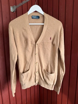 Beige kofta från Polo Ralph Lauren - Klassisk beige kofta från Polo Ralph Lauren med v-ringning, knäppning framtill och två fickor. Ikoniska loggan broderad på bröstet i vinrött. Mjuk och tunn stickning, perfekt för lager på lager. Passar dig som gillar stilrena och tidlösa plagg.