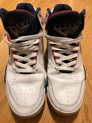 Nike Air Flight vita sneakers - Nike Air Flight sneakers i vitt läder med svarta och röda detaljer. Klassisk hög modell med svart swoosh på sidan, perforeringar på tån och Flight-logga på plösen. Fodrad insida i mörkblått och röda accenter vid snörningen. Perfekta för dig som gillar retrostil.