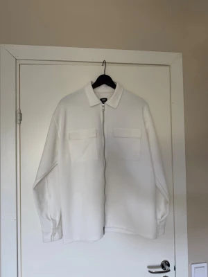 Vit overshirt - Stilren vit overshirt med två stora bröstfickor och dragkedja framtill. Overshirten har klassisk krage, lång ärm och en loose passform. Perfekt att slänga över en t-shirt för en clean look. Använd 2 gånger så den är som ny!