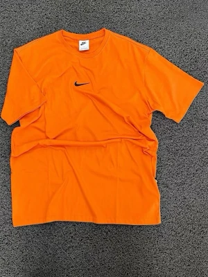 Orange Nike t-shirt med logga - Snygg orange t-shirt från Nike med klassisk svart swoosh-logga på bröstet. T-shirten har rund halsringning och korta ärmar, perfekt för en sportig och avslappnad stil.