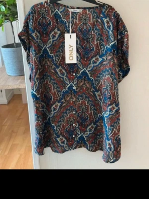 Mönstrad blus från ONLY - Snygg blus från ONLY med färgstarkt paisleymönster i blått, rött och beige. Blusen har korta ärmar och knappar framtill. Tillverkad i ett lätt och luftigt material som passar perfekt till sommaren.