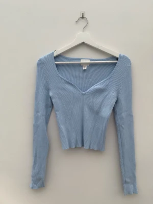 Ljusblå ribbad croppad topp - Säljer en ljusblå ribbad topp från H&M med v-ringad hals och långärmad design. Toppen är croppad och har en slim passform som framhäver siluetten. Perfekt att matcha med jeans eller kjol för en snygg look.