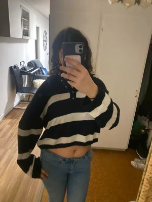 Svartvit randig cropped stickad tröja - Säljer en knappast använd svart och vit stickad tröja med breda horisontella ränder. Tröjan är cropped och har långa, vida ärmar samt knappar vid halsen. Perfekt för dig som gillar en avslappnad och trendig stil.