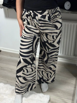 Zebramönstrade vida leggings - Säljer ett par zebramönstrade leggings med svart och beige mönster. De har bred resår i midjan och vida ben för en avslappnad och trendig look. Materialet ser ut att vara mjukt och följsamt, perfekt för dig som gillar statement-plagg.