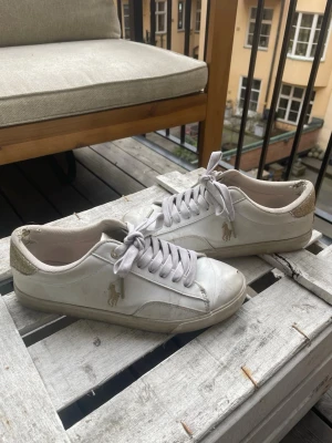 Vita/guld sneakers från Polo Ralph Lauren - Ralph lauren sneakers dam i strl 37 med fina glitter detaljer! Ganska slitna, finns ett litet hål på ena skon därav priset. Köpta för 900 för något år sedan. 