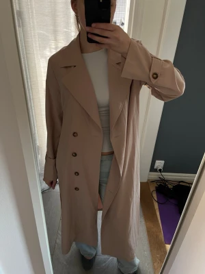Beige trenchcoat  - Stilren beige lång trenchcoat med dubbelknäppning och stora slag. Tunn modell- perfekt för våren 