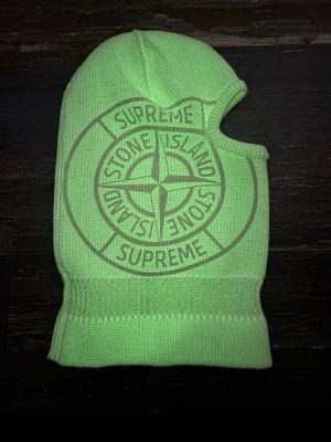 Supreme x Stone Island balaclava - Stickad balaclava i limegrönt och vitt med stor Supreme x Stone Island-logga framtill. Tight passform med öppning för ansiktet och ribbad kant nedtill. Perfekt för att sticka ut i vinterkylan.