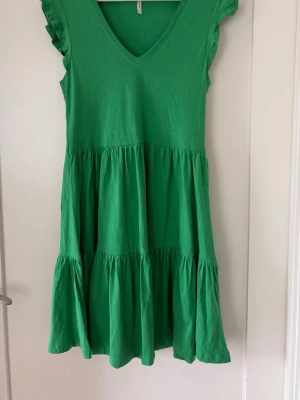 Grön volangklänning med v-ringning från Only - En grön klänning med v-ringad hals, volangdetaljer på axlarna och flera lager i kjolen för en luftig känsla. Klänningen är kort och har en avslappnad passform, perfekt för varma dagar. Tillverkad i mjuk bomull. Längd 90 cm. Är XS men tycker den passar S också.