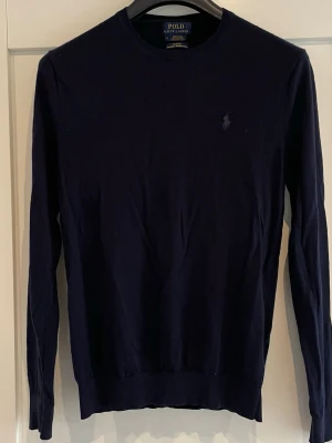 Mörkblå stickad tröja Polo Ralph Lauren - Snygg mörkblå stickad tröja från Polo Ralph Lauren med rund halsringning och diskret broderad logga på bröstet. Tröjan har långa ärmar och ribbade muddar vid ärmslut och nederkant. Perfekt för en stilren look.