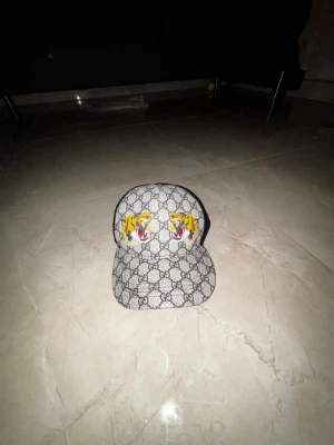  keps med tigertryck - Snygg keps med klassiskt GG-mönster och två färgglada tigerhuvuden framtill. Kepsen har svart mesh på baksidan för extra ventilation och justerbar passform. Perfekt för dig som vill sticka ut med en lyxig accessoar.