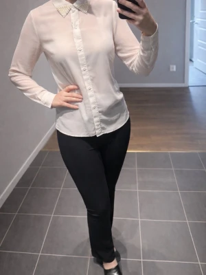 Vit blus med pärlkrage från veromoda  - Stilren vit blus från veromoda med lång ärm och genomskinligt material. Kragen är dekorerad med pärlor för en extra touch. Perfekt att matcha med både jeans och kjol. Klassisk knäppning framtill och smal passform.