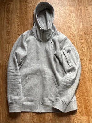 Grå hoodie från Pelle P - Snygg grå hoodie från Pelle P med dragkedja, broderad logga på bröstet och patch på ärmen. Mjuk bomullsblandning, två fickor fram och justerbar huva med snören. Perfekt för en avslappnad och sportig stil.