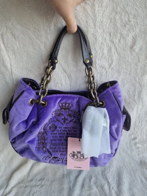 Juicy couture fairytale Lila sammetshandväska Juicy Couture - Superfin lila handväska från Juicy Couture i mjuk sammet med dekorativa kedjor och svarta handtag i skinn. Väskan har broderad text och krona på framsidan samt detaljer i guld. Perfekt för dig som vill sticka ut med en färgglad accessoar.