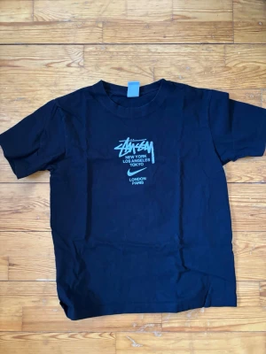 Svart Stüssy t-shirt med tryck - Svart t-shirt från Stüssy med ikoniskt tryck på bröstet i vitt och blått. Klassisk passform med rund hals och korta ärmar. Trycket listar städer som New York, Los Angeles, Tokyo, London och Paris. Perfekt för dig som gillar streetwear.