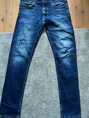 Mörkblå slim fit jeans med slitningar - Snygga mörkblå jeans i slim fit-modell med slitna detaljer på knäna och klassiska fem fickor. Jeansen har en något tvättad look och raka ben. Perfekta för en avslappnad och trendig stil.
