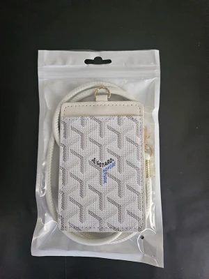 Vit korthållare med snodd - Snygg vit korthållare med klassiskt mönster och blå logga framtill. Kommer med matchande vit axelrem och guldfärgade metalldetaljer. Perfekt för kort och ID med genomskinlig ficka på baksidan. Tillverkad i canvas och läder.