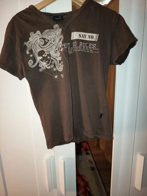 Brun t-shirt med tryck och v-ringning - Cool brun t-shirt med v-ringning och vit grafisk print framtill med texten 'Say No! Accept No Rules!'. Trycket har ett konstnärligt motiv av ett ansikte och blommor. Perfekt för dig som gillar unika statement-plagg.