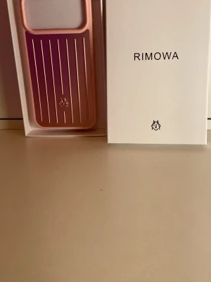 Rosa metalliskt mobilskal från Rimowa - Snyggt mobilskal från Rimowa i metallisk rosa färg med vertikala linjer och logotyp på baksidan. Skalet har en stilren och modern design, tillverkat i tåligt material som ger exklusiv känsla och bra skydd till din mobil.