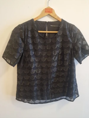 Svart glittrig topp från ONLY - Snygg svart topp från ONLY med glittrigt fjällmönster och rund halsringning. Toppen har korta ärmar och är tillverkad i ett lätt, transparent material som ger en cool och trendig look.