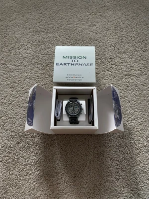 Omega x Swatch MoonSwatch Mission To Earthphase  - Snygg Omega x Swatch MoonSwatch från Bioceramic MoonSwatch Collection, modell Mission to Earth. Klockan har en rund urtavla i silver med svarta detaljer och svart textilband med Omega-logga. Perfekt för dig som gillar stilrena och unika accessoarer.