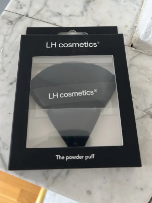 Svart powder puff från LH cosmetics - En svart powder puff från LH cosmetics, perfekt för att applicera puder jämnt över ansiktet. Puffen har en halvmåneformad design och kommer i en snygg förpackningen . Tillverkad i mjukt material som känns skonsamt mot huden.