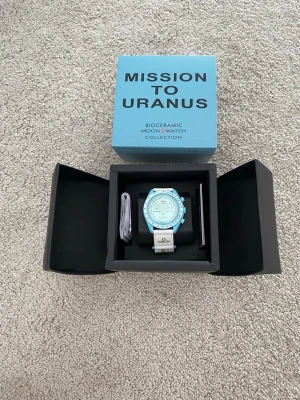 Omega x Swatch MoonSwatch Uranus - Snygg Omega x Swatch MoonSwatch Mission to Uranus med ljusblå boett och vit textilrem. Klockan har en unik biokeramisk design och flera urtavlor med tachymeter. Perfekt för dig som gillar coola och trendiga accessoarer.