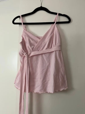Ljusrosa omlottopp med knytband - Söt ljusrosa omlottopp med smala axelband och knytband i midjan. Toppen har v-ringad omlottdesign och är tillverkad i mjuk bomull, perfekt för varma dagar. Lätt utsvängd passform och fina detaljer både fram och bak.