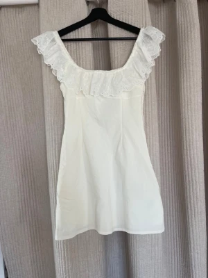 Vit kort klänning med spetsvolang - Supersöt vit kort klänning med bred spetsvolang runt axlarna och en liten rosa rosett framtill. Klänningen har en feminin och romantisk stil, är ärmlös och har en lätt A-linjeform. Perfekt för sommarens alla tillfällen! Passar även till studenten🥰