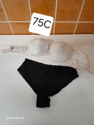 Vit spets-bh och svarta trosor set - Säljer ett set med en vit bh i spets med vadderade kupor och justerbara axelband, storlek 75C, samt ett par svarta trosor med diskret spetskant. Perfekt för dig som vill ha ett snyggt och bekvämt underklädesset.