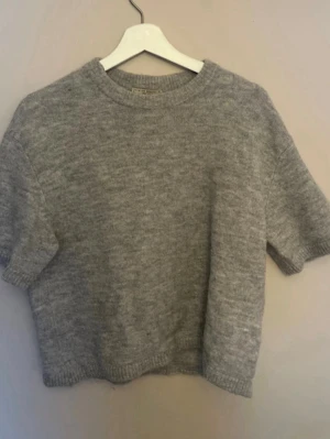 Grå stickad t-shirttröja - Kortärmad grå stickad tröja med rund hals och ribbade kanter. Perfekt för lager-på-lager och enkel att styla till olika outfits. Mjuk och skön känsla.