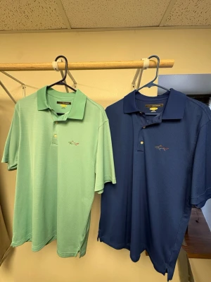 Greg Norman pikétröjor i blått och mint - Två snygga pikétröjor från Greg Norman, en i mintgrön och en i mörkblå. Båda har klassisk krage, knappar framtill och ett broderat hajmotiv på bröstet. Materialet är polyester och tröjorna har korta ärmar. Perfekt för dig som gillar sportig stil. En för 300kr båda för 500kr